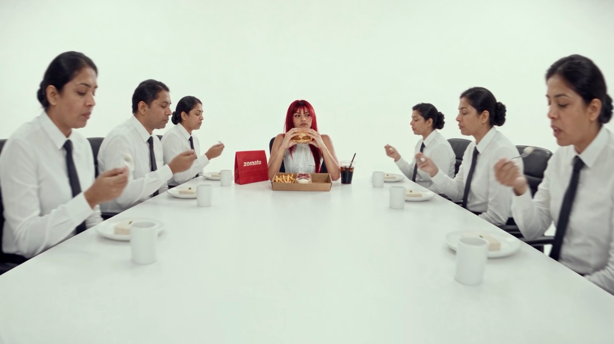 A woman with red hair takes a bite of a burger at a long white table of office workers in matching uniforms