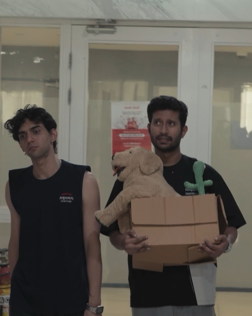 Manav walks through the lobby beside Rohan, who carries the box with the teddy bear and the cactus