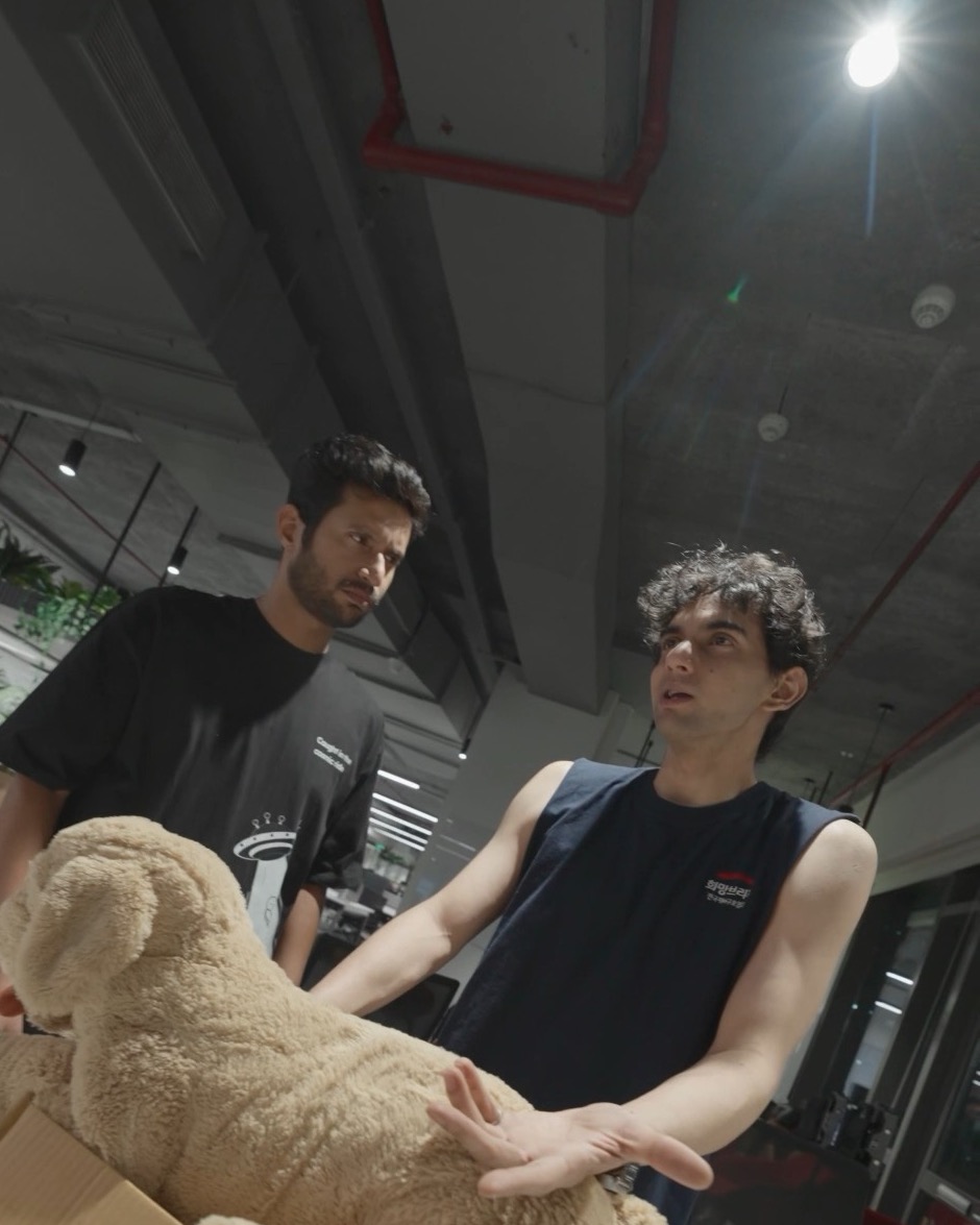 At the desk, Manav explains his plan while Rohan listens, a teddy bear in the foreground