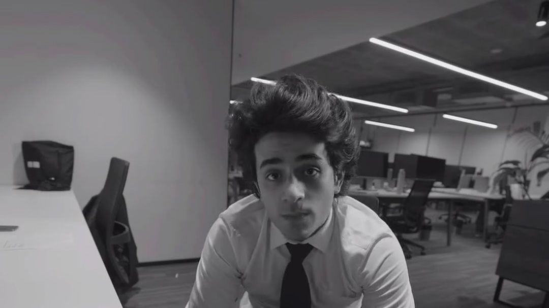 In black and white, a young man in a shirt and tie leans into the camera in an office